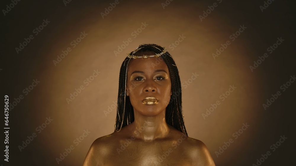 Portrait of a seminude African American woman in the style of Cleopatra in the studio on a brown background with circular light. A woman with creative golden makeup, golden skin, jewelry in her hair. Portrait of a seminude African American woman in the style of Cleopatra in the studio on a brown background with circular light. A woman with creative golden makeup, golden skin, jewelry in her hair.