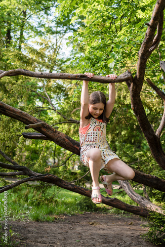 The young girl in a summer jumpsuit is climbing a tree during the summer.
