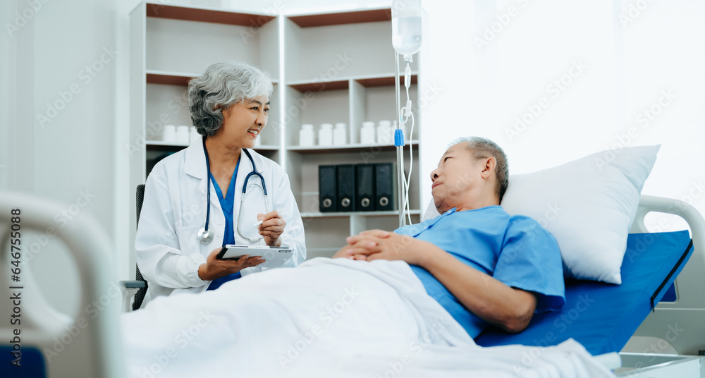 Asian doctor in white suit take notes while discussing and Asian elderly, man patient who lying on bed with receiving saline solution in hospital or clinic. Asian doctor in white suit take notes while discussing and Asian elderly, man patient who lying on bed with receiving saline solution in hospital or clinic.