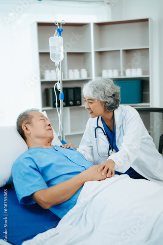 Asian doctor in white suit take notes while discussing and Asian elderly, man patient who lying on bed with receiving saline solution in hospital or clinic.