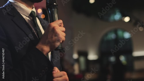 Crop of male event planner speaking in microphone and entertaining public on wedding in restaurant. Handsome gentleman actively gesturing hand while giving speech and proposing toast.