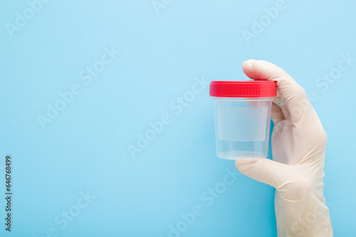 Doctor hand in white rubber protective glove holding transparent plastic container for urine or other analysis test on light blue background. Pastel color. Closeup. Empty place for text. Front view.