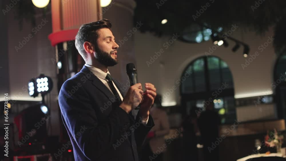 Young caucasian wedding host dressed in black suit taking word and speaking in front of audience in fancy restaurant. Bearded man with microphone in hands expressing best wishes to newlyweds. Young caucasian wedding host dressed in black suit taking word and speaking in front of audience in fancy restaurant. Bearded man with microphone in hands expressing best wishes to newlyweds.