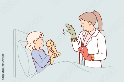 Caring woman pediatrician stands near bed of little girl in hospital and tries to make child laugh
