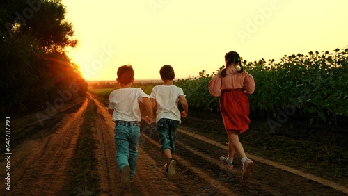 Children play, run through field of sunflowers, friends run at sunset. Happy childhood dream concept. Happy boy girl running, playing together in nature. Family, kids, weekend outdoors. Children walk
