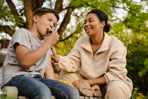 Little boy eating candy bar while sitting with his mother in park