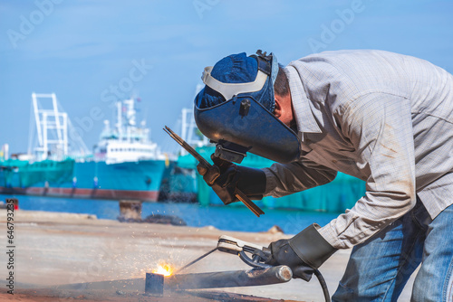 Welder with safety equipment using arc welding machine to welding galvanized steel pipe in shipyard area at harbor