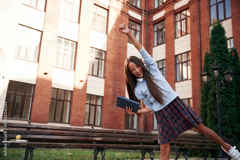 Cheerful, celebrating success. School girl in uniform is outdoors near the building Cheerful, celebrating success. School girl in uniform is outdoors near the building