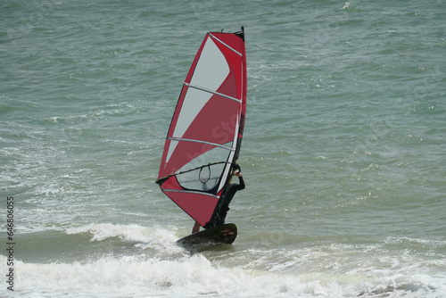 Windsurfer in action on a board with a red and white sail on white waves of the sea.