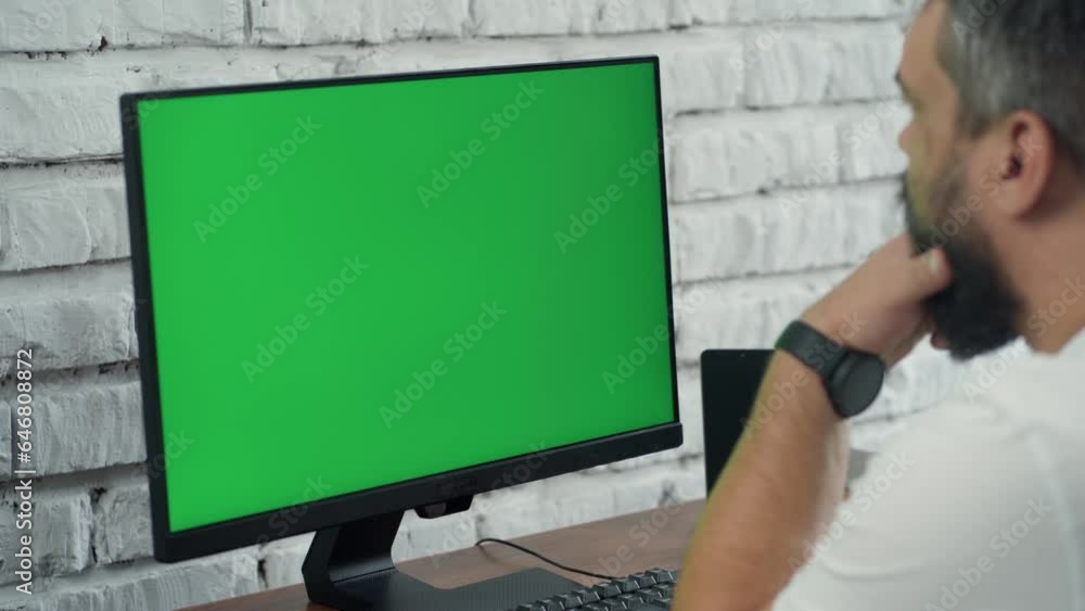 Bearded Middle-Aged Man Working at Desktop Computer With Green Mock Up Screen. Man Looking at Display. Interior- Modern Office With White Brick Wall Bearded Middle-Aged Man Working at Desktop Computer With Green Mock Up Screen. Man Looking at Display. Interior- Modern Office With White Brick Wall
