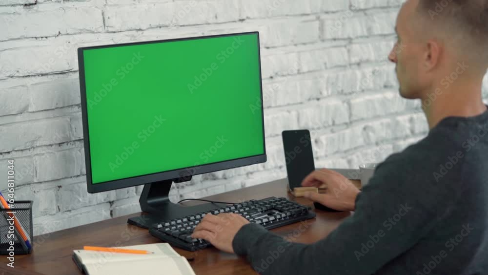 Man Looks at Computer Green Screen and Writes Something in Notebook. Young Man Works In Modern Office With White Brick Wall Man Looks at Computer Green Screen and Writes Something in Notebook. Young Man Works In Modern Office With White Brick Wall