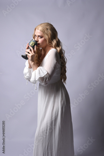 Close up portrait of beautiful blonde model wearing elegant white halloween gown, a historical fantasy character. Holding wine goblet, isolated on audio background.