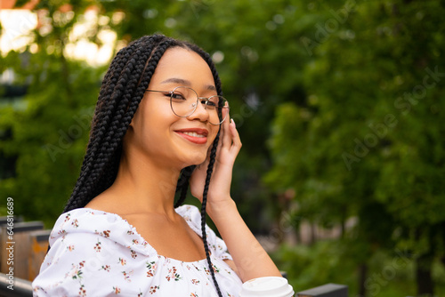 African young attractive woman in glasses stands on a bridge with a paper cup of coffee on the street during the day in the park in summer and looks at the camera