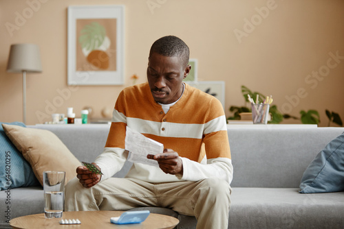 Portrait of adult African American man reading instructions for prescription pills while sitting on couch at home, copy space