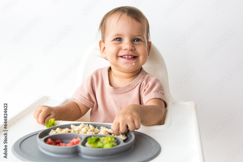 beautiful baby 1 year old sitting in a highchair and eating complementary foods, smiling and looking at the camera on a white background beautiful baby 1 year old sitting in a highchair and eating complementary foods, smiling and looking at the camera on a white background