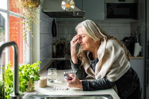 Senior woman taking medication for a migraine in the kitchen at home