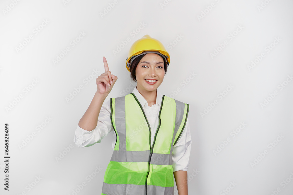 A smiling Asian woman labor wearing safety helmet and vest, pointing to copy space above her, isolated by white background. Labor's day concept.