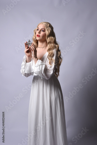 Close up portrait of beautiful blonde model wearing elegant white halloween gown, a historical fantasy character. Holding crucifix cross necklace, isolated on studio background.
