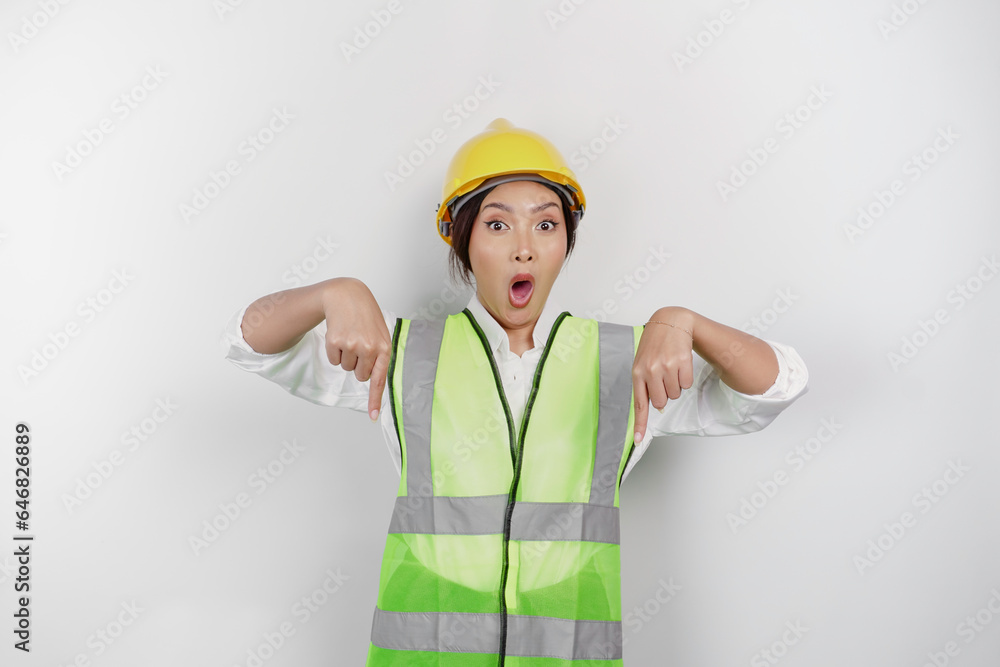 A shocked Asian woman labor wearing safety helmet and vest, pointing to copy space below her, isolated by white background. Labor's day concept.