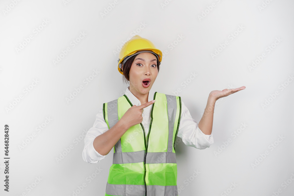 A shocked Asian woman labor wearing safety helmet and vest, pointing to copy space beside her, isolated by white background. Labor's day concept.