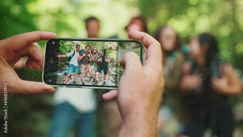 Guy taking pictures of his cheerful backpacking friends on cell phone near mountain river in forest