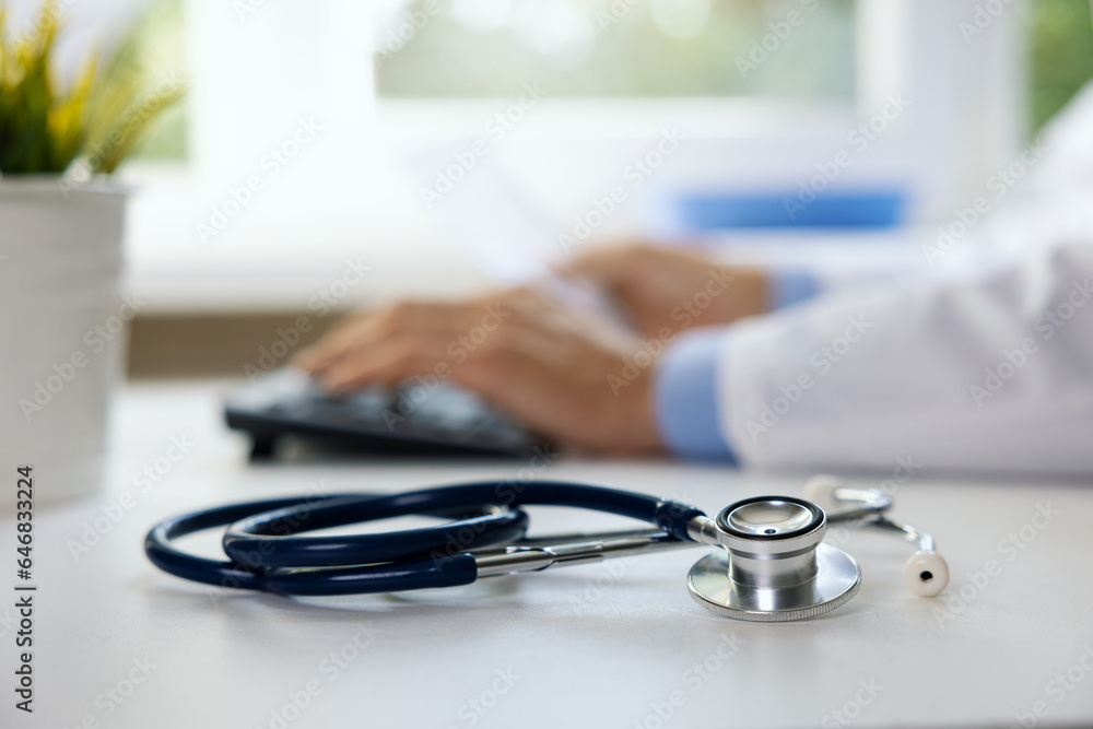 stethoscope on the table in clinic office. doctor working on computer in background stethoscope on the table in clinic office. doctor working on computer in background