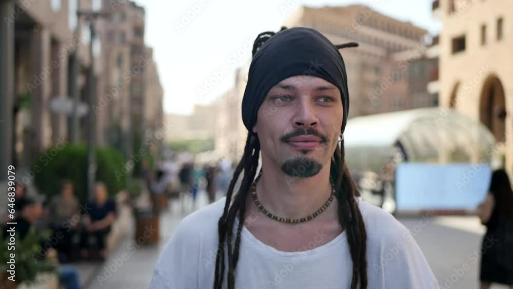 A lonely young handsome man with long dreadlocks and beads walks along the sidewalk of a city street in slow motion and blurred bokeh. Cute walking man with depth of field A lonely young handsome man with long dreadlocks and beads walks along the sidewalk of a city street in slow motion and blurred bokeh. Cute walking man with depth of field