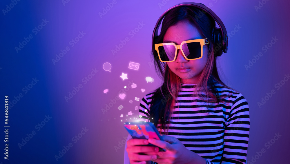 Young Asian teen wearing sunglasses and headphones using smartphone communicating with friends on social media in a room with blue and red neon lights. - obrazy, fototapety, plakaty 