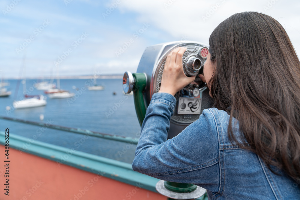 rear view of asian Taiwanese female traveler using sightseeing binoculars to watch sailing boats on the sea at Old Fisherman's Wharf in California usa