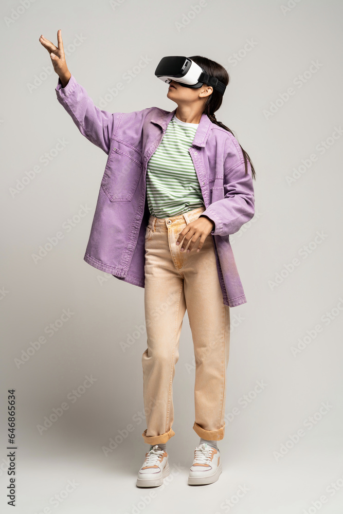 Teenage woman in virtual goggles plays video game points index finger on copy space on gray background. Teen girl looks at virtual world in VR helmet. Cyberspace, advertisement banner, poster concept. Teenage woman in virtual goggles plays video game points index finger on copy space on gray background. Teen girl looks at virtual world in VR helmet. Cyberspace, advertisement banner, poster concept.