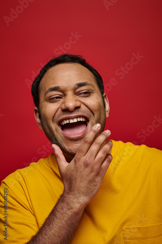 positive emotion, excited indian man in yellow t-shirt laughing with opened mouth on red background © LIGHTFIELD STUDIOS