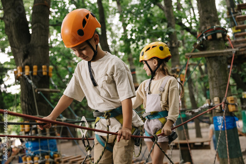 A little smiling girl and boy climbs a rope harness in a summer rope park pass obstacles