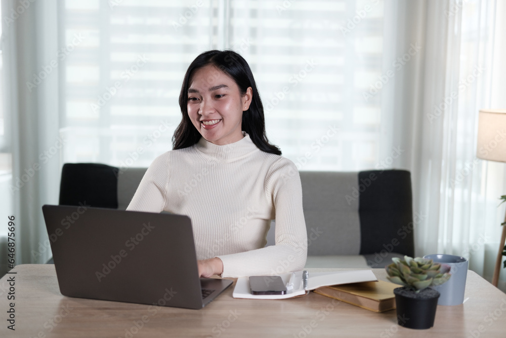 An attractive and happy young Asian woman is working on her laptop at a table in her living room. Working from home, freelance, online meeting