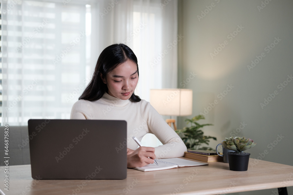An attractive and happy young Asian woman is working on her laptop at a table in her living room. Working from home, freelance, online meeting An attractive and happy young Asian woman is working on her laptop at a table in her living room. Working from home, freelance, online meeting