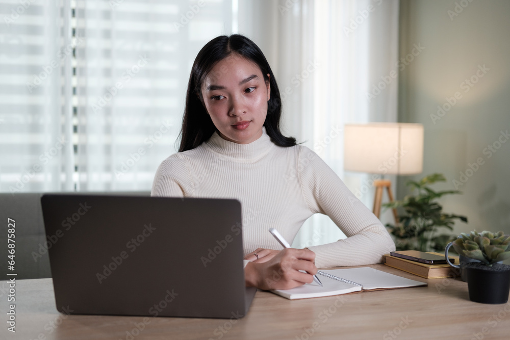 An attractive and happy young Asian woman is working on her laptop at a table in her living room. Working from home, freelance, online meeting An attractive and happy young Asian woman is working on her laptop at a table in her living room. Working from home, freelance, online meeting