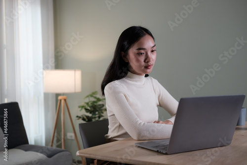 An attractive and happy young Asian woman is working on her laptop at a table in her living room. Working from home, freelance, online meeting © PaeGAG