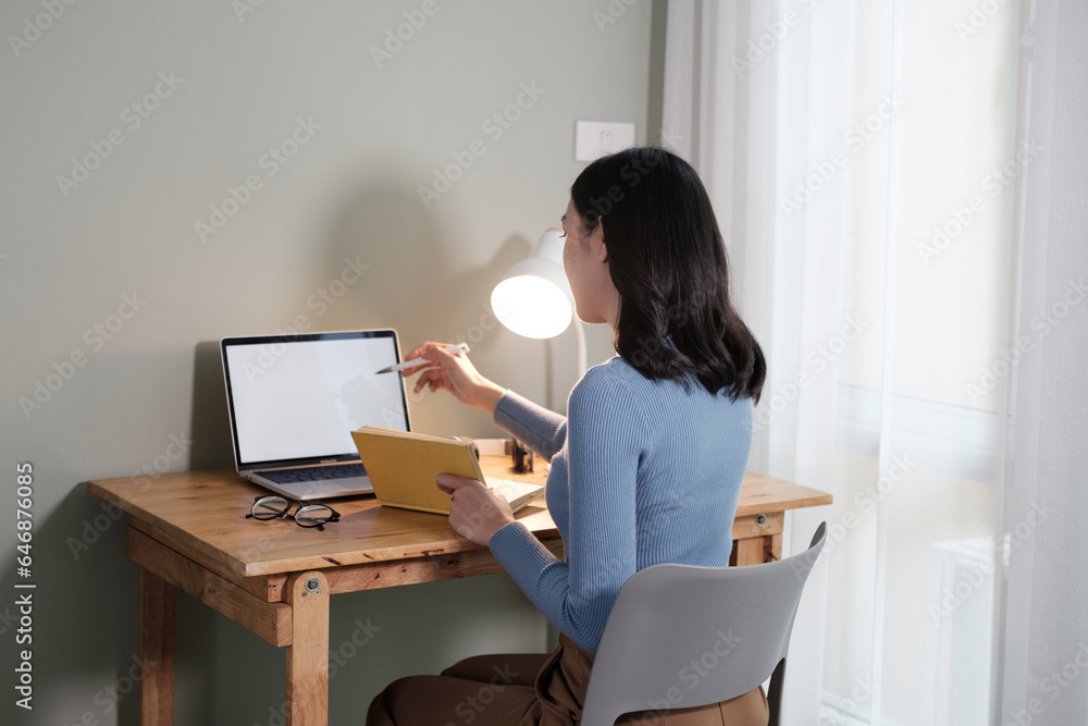 An attractive and happy young Asian woman is working on her laptop at a table in her living room. Working from home, freelance, online meeting An attractive and happy young Asian woman is working on her laptop at a table in her living room. Working from home, freelance, online meeting