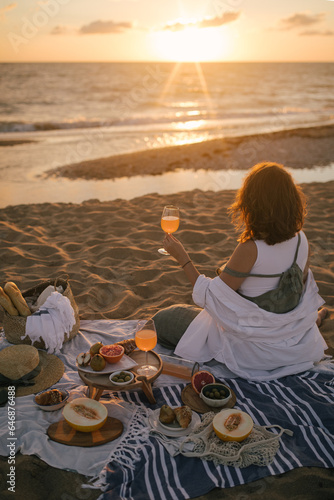 Young woman having beautiful tasty picnic with lemonade, fresh fruits and croissants on a beach at sunset.