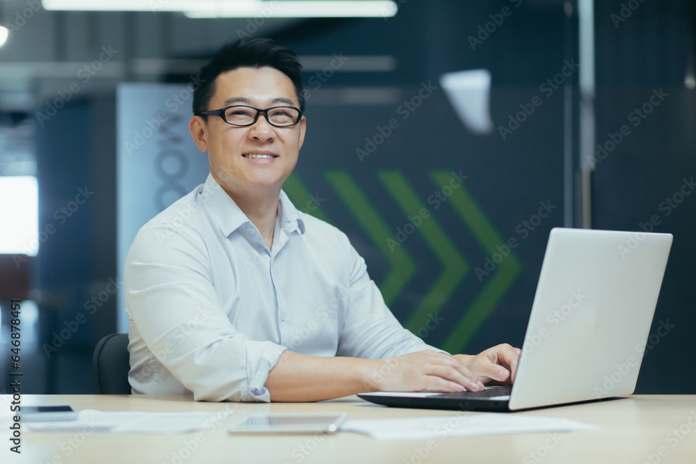 Portrait of a young Asian male architect, engineer in glasses working in the office, sitting at the table at the laptop and smiling at the camera. Portrait of a young Asian male architect, engineer in glasses working in the office, sitting at the table at the laptop and smiling at the camera.