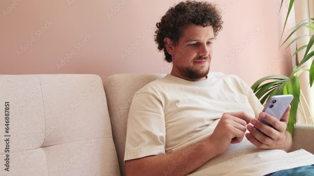 Guy sits on the sofa in cozy living room and scrolls across the phone screen Guy sits on the sofa in cozy living room and scrolls across the phone screen