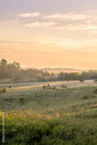 A morning landscape of misty fields and hills with green trees and bushes during the golden hour, against a backdrop of bright sunny sky with clouds.