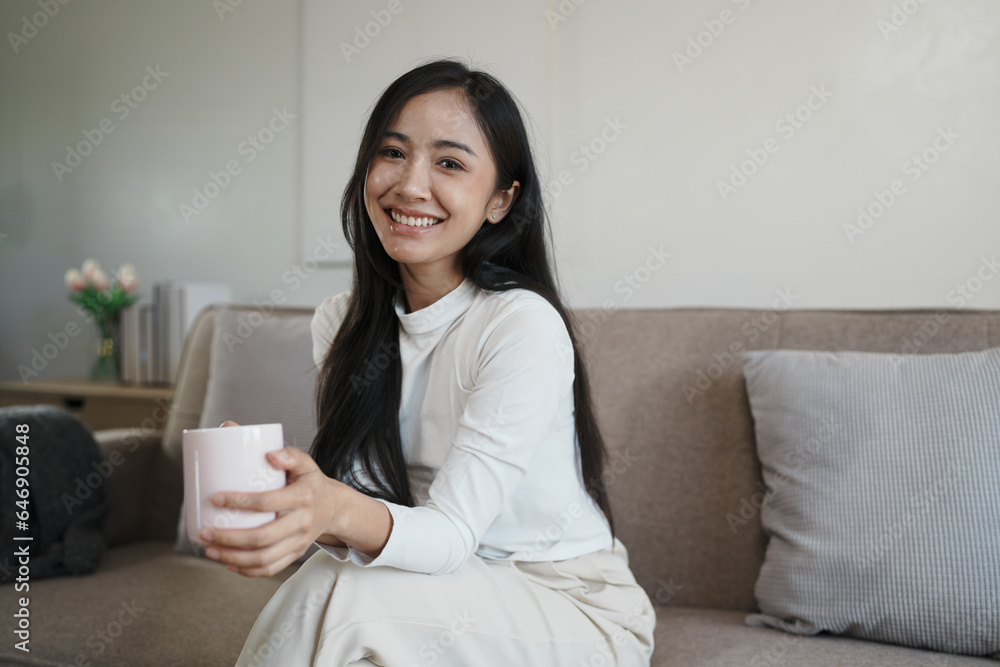 Happy young asian woman drinking coffee relaxing on sofa at home. Smiling female enjoying resting sitting on couch in modern living room. Happy young asian woman drinking coffee relaxing on sofa at home. Smiling female enjoying resting sitting on couch in modern living room.