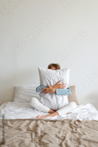 Healthy sleep, dream concept. Teenager boy sitting on bed, embracing pillow, minimalist nordic bedroom interior, lifestyle