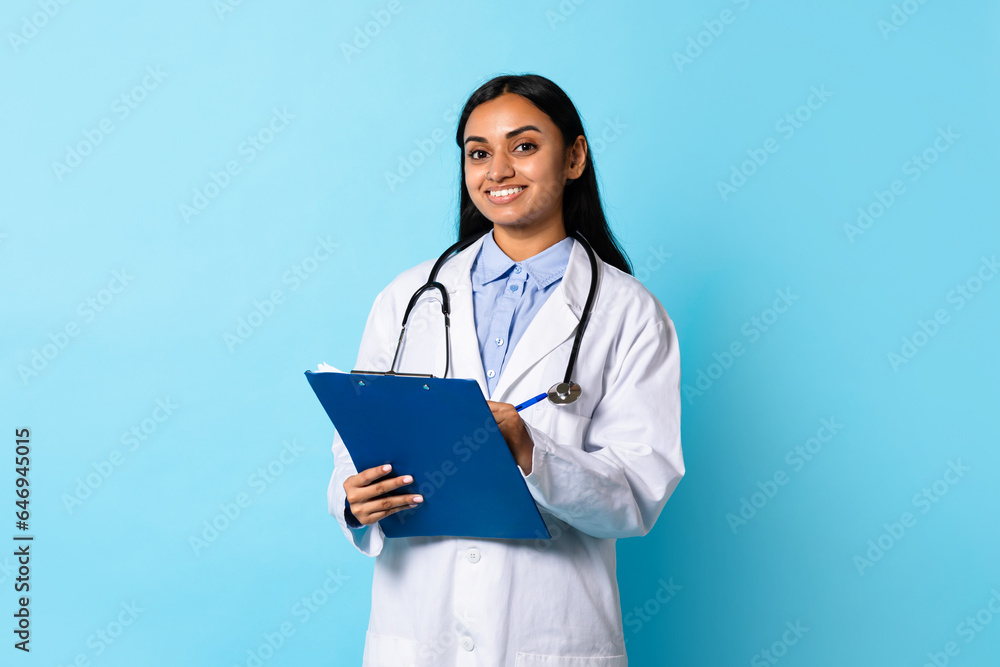 Indian Doctor Lady In Coat Posing With Clipboard, Blue Background