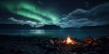 Burning campfire on the beach. Fjord landscape with snow capped mountains. Night sky with stars and northern lights.