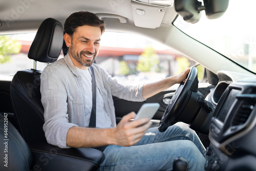 Happy handsome millennial guy driver using smartphone while driving car