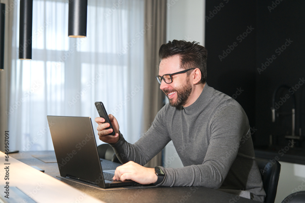 Man is using cell mobile phone at home while working on laptop computer and smile Man is using cell mobile phone at home while working on laptop computer and smile