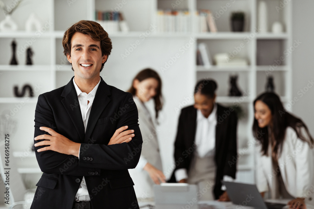 Happy european businessman in suit standing with folded arms in front of his colleagues, posing in office during meeting Happy european businessman in suit standing with folded arms in front of his colleagues, posing in office during meeting
