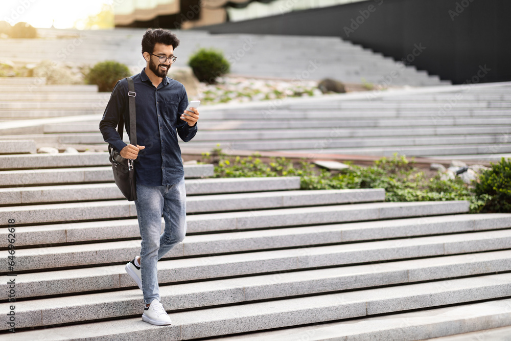 Cheerful indian guy going down stairs outdoors, using cell phone Stock