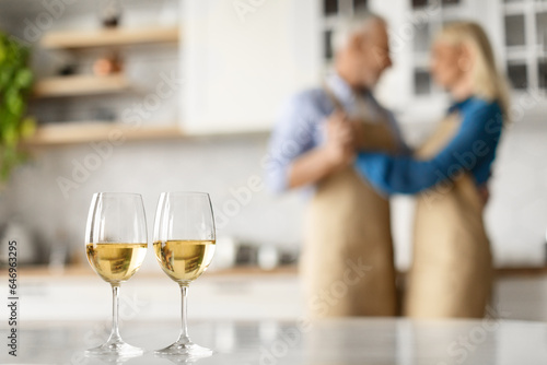 Selective focus on two glasses of white wine on kitchen table over dancing senior couple
